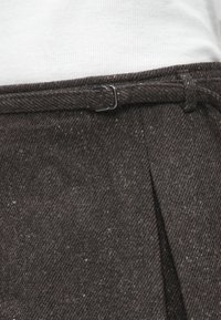 Λευκό ύφασμα κρυμμένο μέσα σε σκούρα καφέ υφασμάτινη φούστα από μάλλινο ύφασμα με λεπτή ζώνη που ταιριάζει και ασημένια αγκράφα στη μέση.