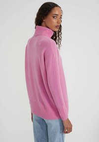 Femme aux cheveux bouclés portant un pull rose ample à col roulé et un jean bleu clair, se retournant par-dessus son épaule devant un fond uni.