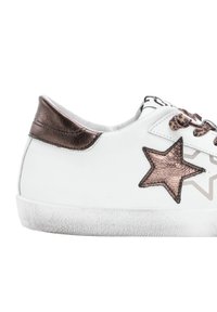Sneaker blanc avec un talon métallique en bronze, une étoile en patch en bronze sur le côté, et des lacets en imprimé léopard, présenté de profil sur un fond blanc.