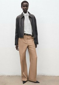 Giacca di pelle nera, maglione grigio con cravatta, pantaloni beige a gamba larga, décolleté nere a punta; design minimalista e palette di colori neutri.