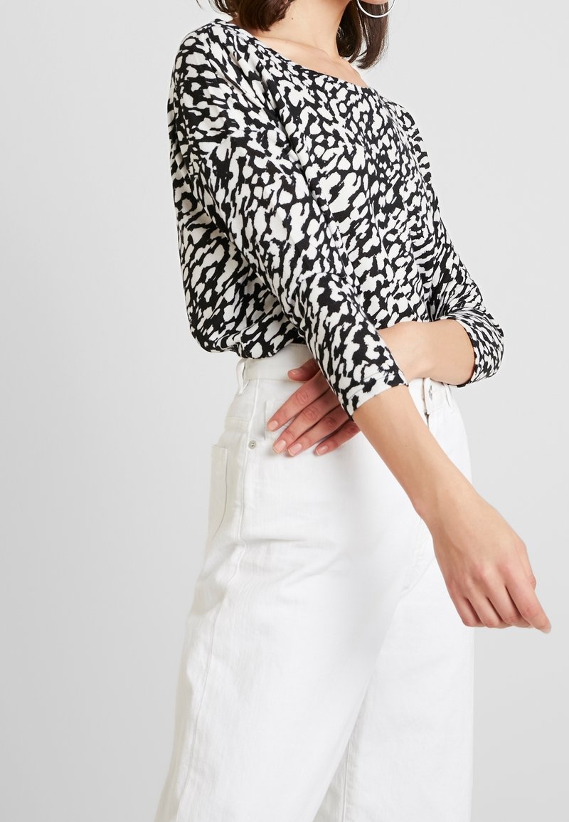 Černobíle vzorované tričko s dlouhým rukávem a lodičkovým výstřihem, v kombinaci s bílými džíny s vysokým pasem, které mají zapínání na knoflík a kapsy.