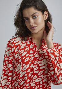Cămașă cu model roșu și alb, cu închidere prin butoni, guler deschis și mâneci lungi. Prezintă un design fluid și o textură moale.