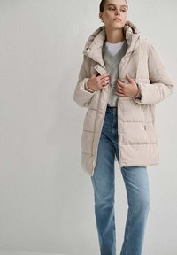 Cappotto imbottito di un beige chiaro con cappuccio, grandi tasche frontali e chiusura con zip. Indossato sopra un maglione grigio e jeans blu. Texture liscia.