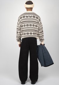 Personne portant un pantalon noir à jambes larges et un pull blanc à motif, tenant une veste matelassée bleue, vue de dos sur un fond uni.