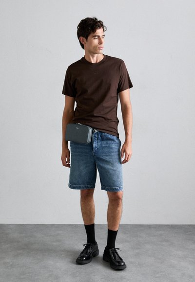 Camiseta de algodón marrón, pantalones cortos de mezclilla azules, bolso cruzado gris, zapatos negros de cordones y calcetines negros en un entorno minimalista.