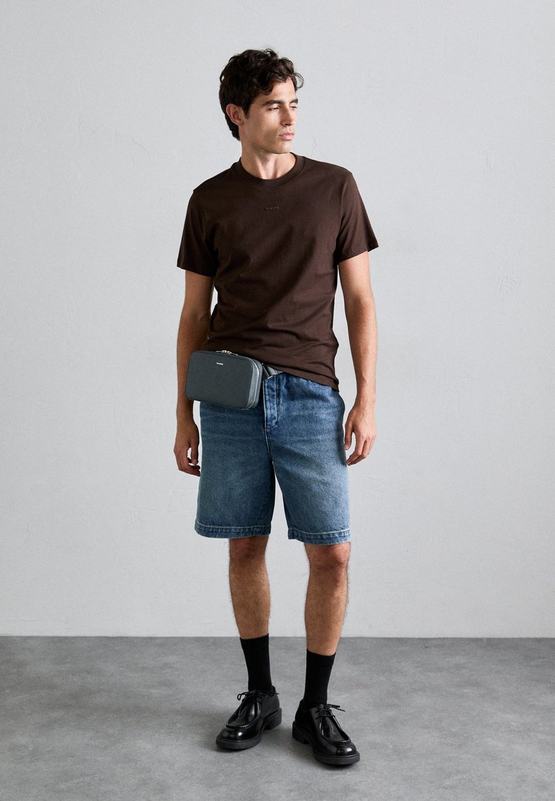 T-shirt in cotone marrone, shorts in denim blu, borsa a tracolla grigia, scarpe nere allacciate e calzini neri in un contesto minimalista.