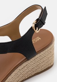 Sandale compensée noire avec une bride texturée, une plateforme couleur cognac et un détail en jute tissé. Comprend une boucle dorée pour un ajustement réglable.