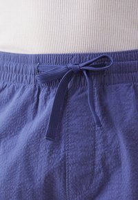 Gros plan sur un pantalon en tissu bleu texturé avec une taille élastique et un cordon noué, porté avec un haut à côtes clair.