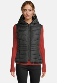 Vrouw in een zwart gewatteerd mouwloos hoodievest over een lange mouw geribbelde rode top en zwarte broek, recht naar voren kijkend met een neutrale uitdrukking.