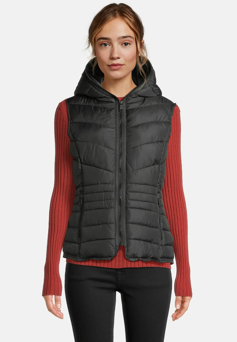 Vrouw in een zwart gewatteerd mouwloos hoodievest over een lange mouw geribbelde rode top en zwarte broek, recht naar voren kijkend met een neutrale uitdrukking.