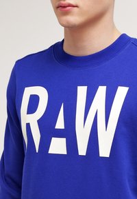 Blå bomullströja med stora vita bokstäver "RAW" och triangulära utskärningar. Ribbad rund halsringning och slät textur.