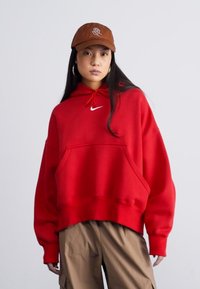 Sweatshirt oversized vermelho com bolso canguru, capuz com cordão e logotipo branco. Usado com calças cargo caqui e um boné castanho com um design bordado.