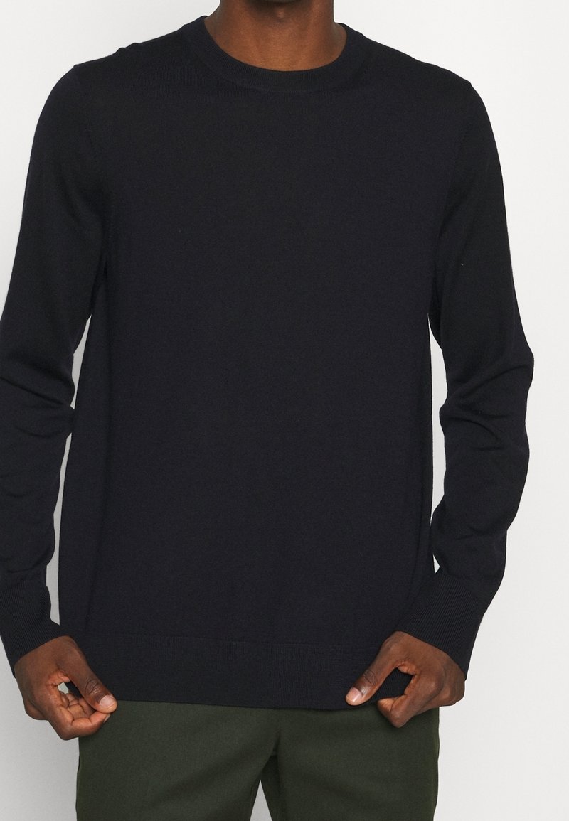 Homme portant un pull noir uni à manches longues avec poignets et ourlet côtelés, posant avec les mains tirant doucement le bas du pull vers le bas.