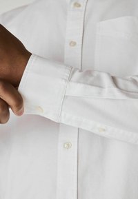 Weißes Hemd mit Knopfleiste aus glattem Material, ausgestattet mit einer einzelnen Brusttasche und geknöpften Ärmelbündchen. Der Ärmelsaum ist teilweise aufgerollt.