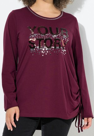 Frau trägt ein langärmeliges bordeauxrotes Oberteil mit schwarzem und silbernem Pailletten-Schriftzug "YOUR STORY" und gerafftem Kordelzugdetail an der Seite.