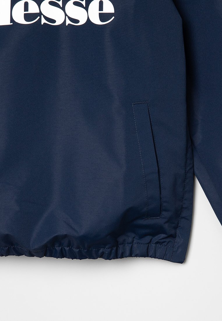 Chaqueta cortavientos de color navy con puños elásticos, que presenta un logo blanco en el pecho y un bolsillo lateral con cremallera oculta. Textura suave.