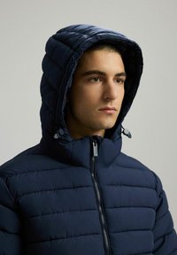 Granatowa pikowana kurtka z wysokim kołnierzem i kapturem. Posiada zamek błyskawiczny z przodu, haftowany wzór oraz regulacje na kapturem za pomocą troków.