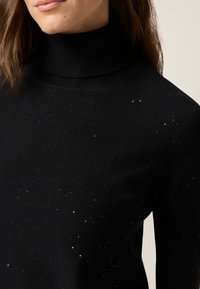 Pull noir à col roulé en tissu doux, présentant une surface texturée avec des détails scintillants argentés dispersés.