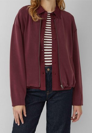 Žena nosící bordó bundu na zip a knoflíky přes pruhované tričko, v kombinaci s tmavě modrými džínami, na jednobarevném světlém pozadí.