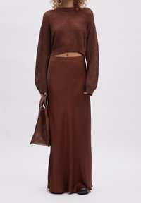 Bruin gebreide cropped trui met een losse fit en lange mouwen, gecombineerd met een bruin satijnen maxi rok. Een bijpassende bruine schoudertas wordt vastgehouden.