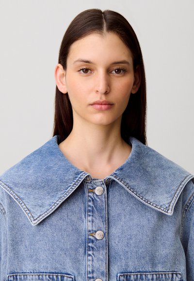Veste en denim bleu clair avec grand col, devant à boutons et deux poches. Présente une finition délavée et des détails de couture visibles.