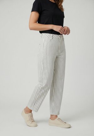 Donna che indossa pantaloni bianchi con strisce nere verticali, maglia a maniche corte nera e sneaker beige con lacci, in piedi contro uno sfondo neutro.