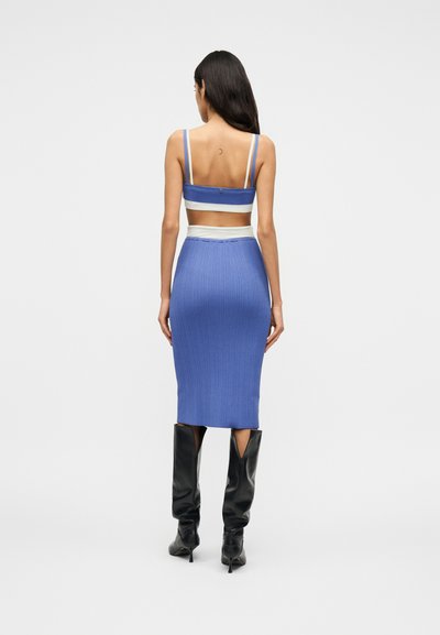 Femme aux longs cheveux noirs portant un débardeur court bleu et blanc, une jupe côtelée assortie et des bottes noires à talons hauts, debout de dos.