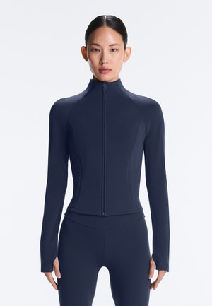 Femme portant une veste zippée bleu marine ajustée avec des trous pour les pouces, associée à un legging bleu marine assorti, debout devant un fond blanc.