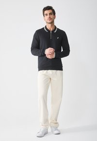 Sort sweatshirt med lynlås, hvid krave og manchetter med stribede detaljer. Sammen med cremede bukser og hvide sko.