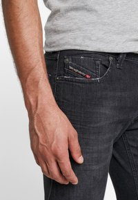 Černé džíny z denimu s opotřebovanými detaily. Obsahují malou přední kapsu s červenobílou značkou Diesel. Detailní záběr ruky opřené o stehno.