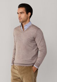 Sveter s V-krkom vyrobený z jemného béžového materiálu, s dlhými rukávmi a rebrovanými manžetami, navrstvený na modrej prúžkovanej košeli s golierom.