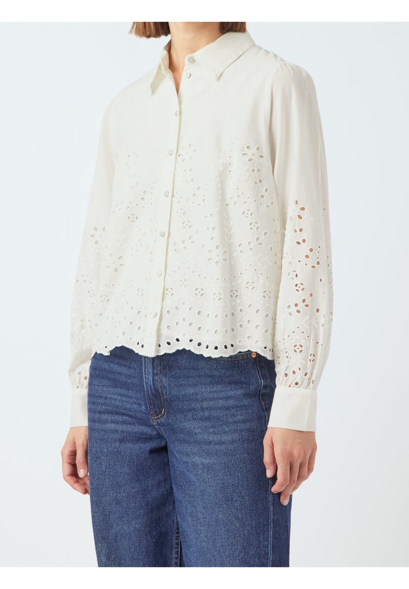 Femme portant un chemisier blanc à manches longues boutonné avec des découpes œillets florales, associé à un jean en denim bleu sur un fond uni.