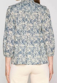 Blommönstrad skjorta med en blå och krämfärgad färgschema, 3/4 ärmar, rynkad detalj vid axlarna och rund halsringning.