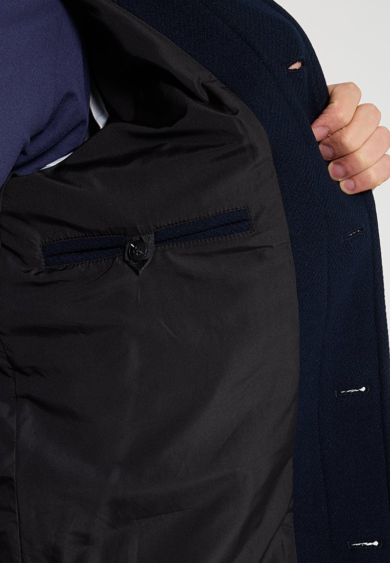 Binnenkant van een marineblauwe colbert met een gladde zwarte voering, voorzien van een klein zakje met knoopsluiting en zichtbare stikdetails.