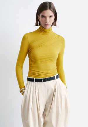 Vrouw die een mosterdgele gerimpelde coltrui draagt, crème wijde geplooide broek met een zwarte riem, zilveren oorringen en armbanden.