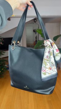 Μαύρη τσάντα tote από δερματίνη με υφή, με δύο λαβές, διακοσμημένη με χρυσά μεταλλικά στοιχεία και φουλάρι με λουλουδένιο μοτίβο δεμένο σε μία από τις λαβές.