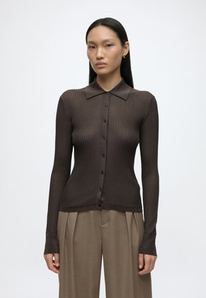 Mujer con cabello negro liso que lleva un top marrón oscuro ajustado con botones y pantalones beige plisados y sueltos sobre un fondo blanco liso.