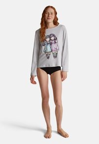 Frau mit langen roten Haaren, die ein hellgraues Langarmshirt mit zwei aufgedruckten Cartoon-Mädchen und schwarze Unterwäsche trägt, barfuß stehend.