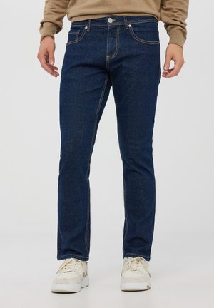 Jeans in denim blu scuro con design a gamba dritta, cuciture visibili, tasche anteriori e chiusura con bottone. Indossati con sneakers bianche.