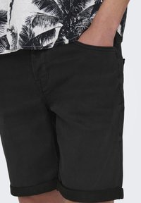 Şorturi din denim negre cu o textură netedă, având un tiv rulat și buzunare laterale. Purtați cu o cămașă albă cu model.