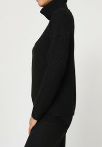Pull à col roulé noir, ample, fabriqué en maille texturée, avec des manches longues et un col haut.