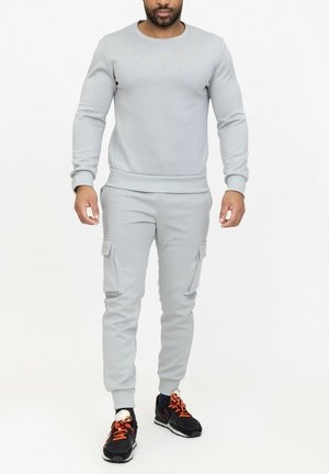 Homme portant un sweat-shirt gris clair et un pantalon de jogging cargo, avec des baskets noires et orange, debout devant un fond blanc.