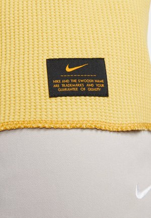 Tessuto giallo testurizzato con un'etichetta nera che presenta il logo Nike. L'etichetta include testo arancione riguardante i marchi e l'assicurazione di qualità.