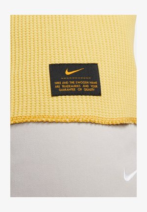 Tessuto giallo testurizzato con un'etichetta nera che presenta il logo Nike. L'etichetta include testo arancione riguardante i marchi e l'assicurazione di qualità.