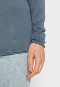Langärmliges blaues Strickoberteil mit gerippten Bündchen, das eine glatte Textur aufweist. Die Hand wird vor hellblauem Denim gezeigt. Der Saum endet auf Hüfthöhe.