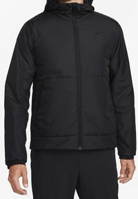 Homme portant une veste zippée noire à capuche avec des poches latérales et un petit logo Nike sur la poitrine, debout devant un fond blanc uni.