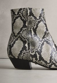 Botină până la gleznă cu imprimeu de șarpe în negru și bej, cu o suprafață texturată, toc conic și finisaj neted, prezentând un model geometric.