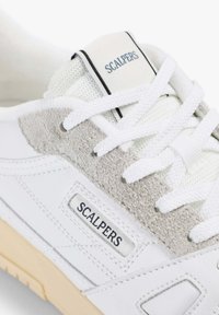 Zapatilla blanca con detalles de ante en gris claro, tela texturizada, lengüeta con marca y cordones planos blancos. Suela de goma visible.