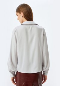 Hellgraue Bluse mit klassischem Kragen, langen Ballonärmeln und einer glatten Textur. Getragen mit einem glänzenden, dunkelroten Rock.