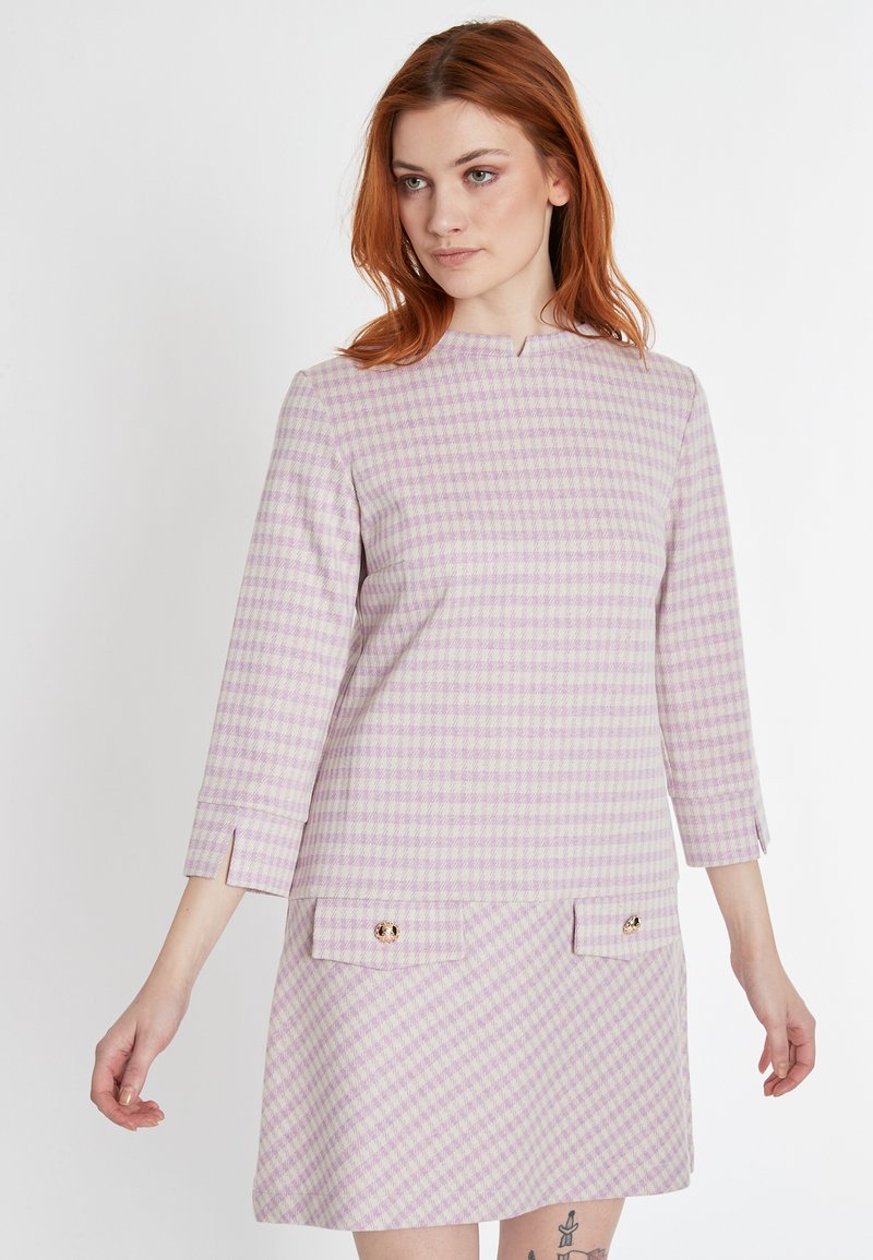 Ein lila-weiß kariertes Kleid mit dreiviertel Ärmeln, Spitzenausschnitt und goldenen Knopfakzenten an den vorderen Taschen. Strukturierter Stoff.
